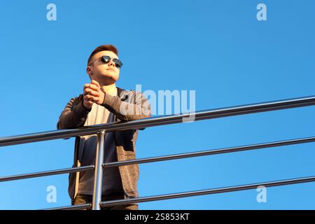 Ein junger Mann in lässigem Outfit und Sonnenbrille lehnt sich an einem modernen Metallgeländer unter klarem blauen Himmel. Er sieht entspannt aus und blickt von der Kamera weg Stockfoto
