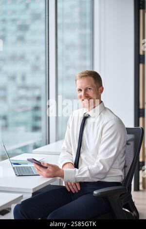 Glücklicher, selbstbewusster junger Geschäftsmann, der im Büro ein Smartphone verwendet. Hochformat. Stockfoto