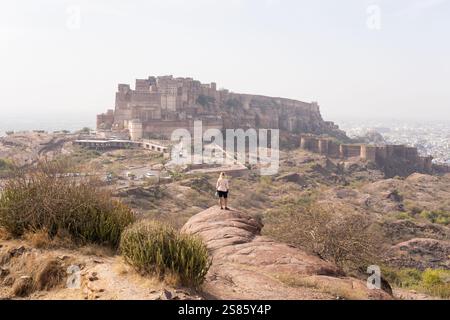 Junge blonde Frau, die den Blick über das Fort Jodhpur genießt, während sie auf dem Felsen in Rajasthan, Indien, steht. Stockfoto