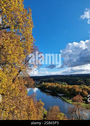 Herbstlandschaft eines Flusses, der sich durch einen Wald schlängelt, der mit lebhaften Herbstfarben gefärbt ist, hellblauer Himmel mit flauschigen Wolken. Ruhige Atmosphäre Stockfoto