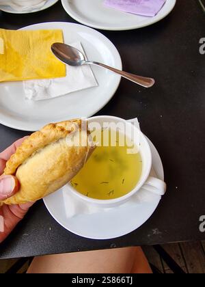 Eine Hand mit einem traditionellen Gebäck (Kibinas) gefüllt mit Hammelfleisch, mit warmer Hühnerbrühe, serviert auf einem dunklen Holztisch, Litauen Stockfoto