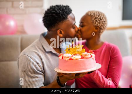 Glückliches junges Paar, das zu Hause den Geburtstag der Frau feiert. Sie halten Kuchen mit Geburtstagskerze und küssen sich. Stockfoto