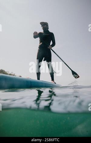 Ein Mann steht auf einem Surfbrett im Wasser. Er trägt einen Neoprenanzug und hält ein Paddel. Das Wasser ist ruhig und der Himmel ist klar Stockfoto