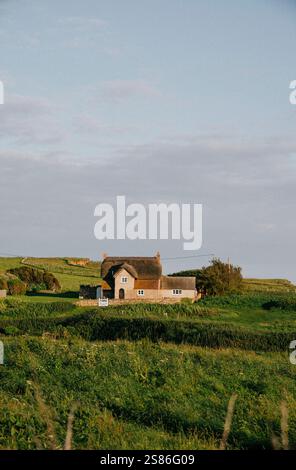 Ein kleines Haus sitzt auf einem Grasfeld. Der Himmel ist bewölkt und die Sonne scheint Stockfoto