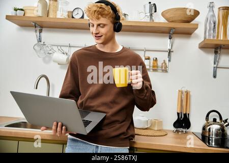 Ein hübscher junger Mann mit roten Haaren arbeitet an seinem Laptop und schlürft Kaffee in seiner stilvollen Küche. Stockfoto