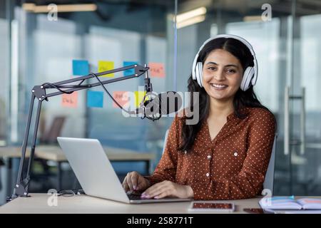 Porträt einer jungen Bloggerin, Influencerin, die lacht und in die Kamera schaut, mit einem professionellen Mikrofon, Kopfhörern und Laptop einen Podcast in einem Studio-Büro aufnimmt. Stockfoto