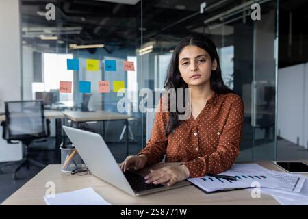 Porträt einer ernsthaften, selbstbewussten Frau bei der Arbeit mit einem Laptop. Geschäftsfrau, die mit Zufriedenheit in die Kamera schaut, im Büro am Arbeitsplatz arbeitet. Stockfoto