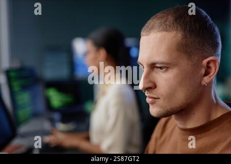 Seitliches Porträt eines konzentrierten männlichen Programmierers mit minimalistischem Silberohrring und Septum, das in das Schreiben von Computercode getaucht ist, am Schreibtisch im modernen IT-Firmenbüro, im Kopierraum Stockfoto