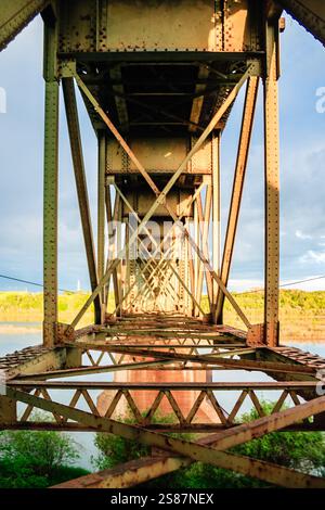 Die Brücke ist alt und verrostet, mit vielen Metallstangen und Drähten. Der Himmel ist bewölkt und das Wasser unter der Brücke ist ruhig Stockfoto