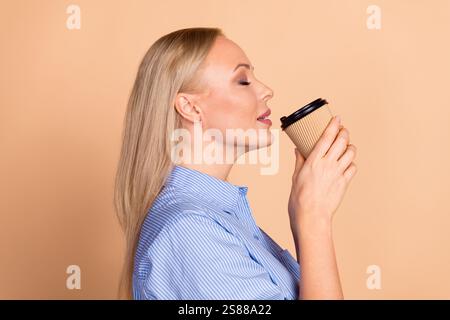 Junge Geschäftsfrau genießt Kaffee in einer Einwegbecher vor beigefarbenem Hintergrund, trägt ein blau gestreiftes Hemd und lässiges Styling. Stockfoto