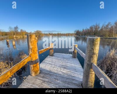 Frostbedeckter Holzsteg über einem gefrorenen Badesee an einem sonnigen Wintertag mit klarem blauen Himmel. Pool Bridge Farm. York. UK Stockfoto
