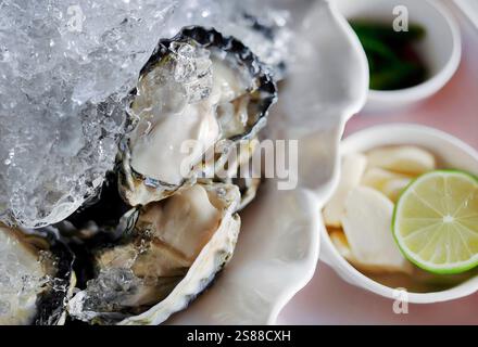 Frische Austern auf der Halbschale auf Eis mit Limettenscheiben und anderen Beilagen, scharfe Thai-Stil, Meeresfrüchte aus dem Meer, lokale Gerichte Stockfoto