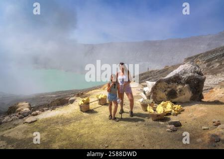 Eine fröhliche Mutter und Tochter stehen zusammen in atemberaubender vulkanischer Landschaft mit Schwefel in Körben, umgeben von Dampf und einzigartigen Formationen Stockfoto