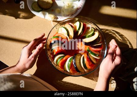Lebendige Gemüsescheiben, einschließlich Zucchini, Tomaten und Auberginen, ordentlich in einer runden Glasschale angeordnet, bereit für die Zubereitung, mit warmem Sonnenlicht Stockfoto