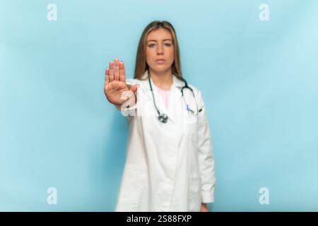 Eine junge Ärztin mit weißem Mantel und Stethoskop, die mit der Hand eine Stoppgeste macht, Verleugnung ausdrückt und Uneinigkeit zeigt, auf blau stehend Stockfoto