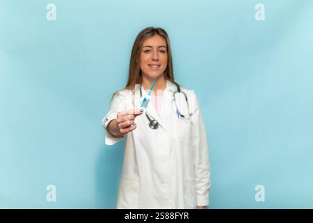 Junge Ärztin mit weißem Mantel und Stethoskop hält und zeigt ein digitales klinisches Thermometer auf hellblauem Hintergrund, lächelnd und lecker Stockfoto