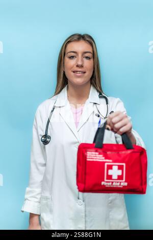 Vertikal freundliche Ärztin mit rotem Verbandskasten, Stethoskop und Labormantel, auf blauem Hintergrund stehend, bietet medizinische Hilfe an Stockfoto