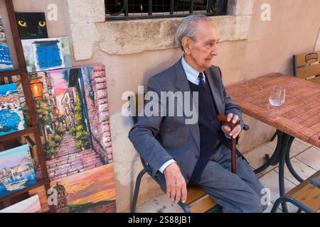 Älterer älterer Mann, der am Café-Tisch mit lokalen Kunstgemälden von lokalen Künstlern sitzt und an Touristen in der ummauerten Altstadt verkauft wird. Dubrovnik, Kroatien. (138) Stockfoto