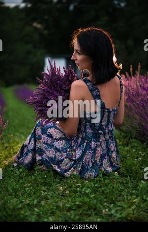 Eine junge Frau in einem gemusterten Sommerkleid sitzt in einem Lavendelfeld. Sie hält einen großen Blumenstrauß der lila Blumen. Die Szene ist eingetaucht Stockfoto