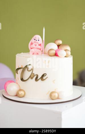 Eine köstlich aussehende Geburtstagskuche, die mit einer niedlichen Hasenfigur, pastellfarbenen Kugeln und einem goldenen „Eins“-Topper verziert ist. Perfekt für einen Feiernden Stockfoto