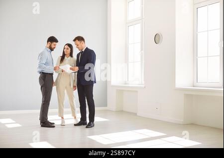 Ein junges Paar, das Dokumente mit einem Immobilienmakler in einem hellen Raum überprüft und über den Hauskauf diskutiert. Stockfoto