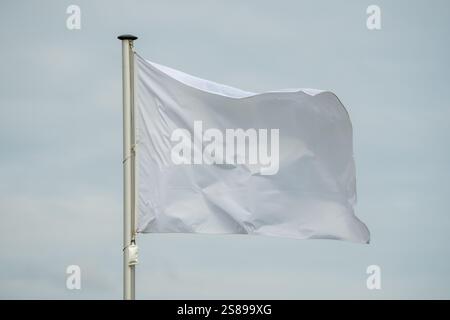 Eine weiße Fahne weht im Wind. Der Fahnenmast ist hoch und die Fahne groß. Der Himmel ist bewölkt und die Sonne ist nicht sichtbar Stockfoto