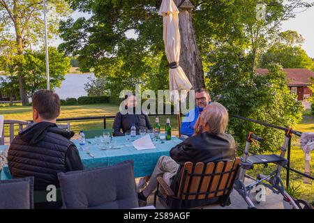 Die Leute sitzen bei Drinks am Tisch im Freien und genießen Gespräche im grünen Garten mit Seeblick an sonnigen Tagen. Schweden. Stockfoto