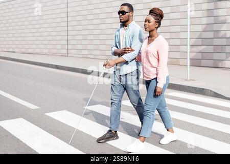 Junge schwarze Frau, die sehbehinderten Millennials mit Stock über die Stadtstraße assistiert. Vision Disability Konzept Stockfoto