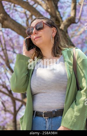Vertikale Aufnahme einer glücklichen Frau in einer grünen Jacke, die unter blühenden Jacarandabäumen am Telefon spricht. Das helle Purp Stockfoto