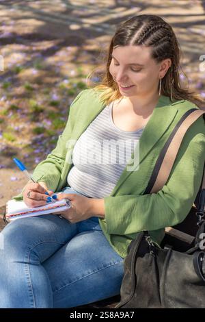 Eine fröhliche junge Frau mit geflochtenen Haaren, von oben nach unten gefangen, in einem Notizbuch, während sie auf einer Bank unter blühenden Bäumen in einer Erbse sitzt Stockfoto
