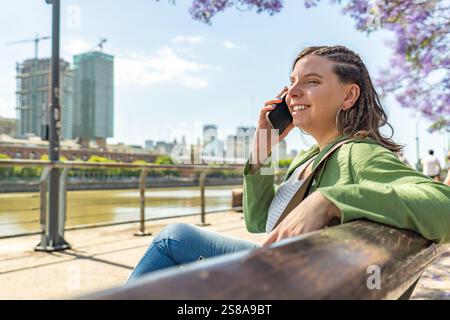 Eine fröhliche Frau, die auf einer Bank am Fluss sitzt und am Telefon spricht, mit modernen Stadtgebäuden und blühenden Bäumen im Hintergrund auf einer sonnigen Sonne Stockfoto