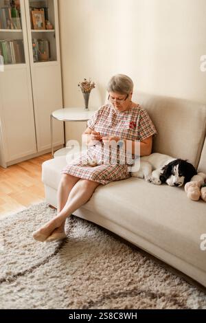 Ältere Dame mit Brille häkeln zu Hause, während ihr weißer Hund neben ihr sitzt. Stockfoto