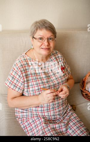 Eine ältere Frau, die zu Hause Brillenhäkeln trägt und auf einem beigefarbenen Sofa sitzt. Stockfoto