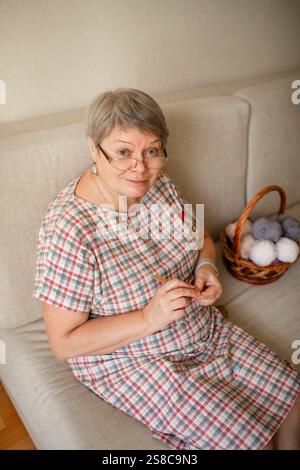 Eine ältere Frau, die zu Hause Brillenhäkeln trägt und auf einem beigefarbenen Sofa sitzt. Stockfoto