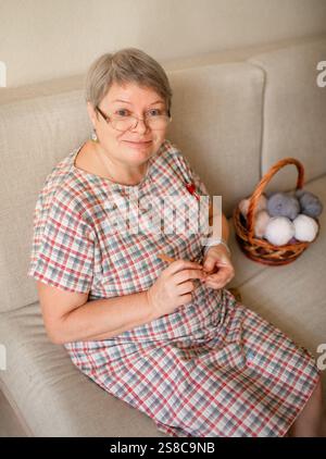 Eine ältere Frau, die zu Hause Brillenhäkeln trägt und auf einem beigefarbenen Sofa sitzt. Stockfoto