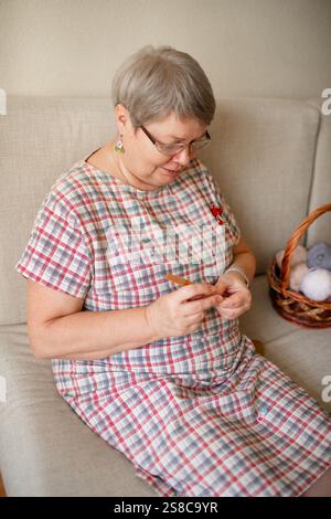 Eine ältere Frau, die zu Hause Brillenhäkeln trägt und auf einem beigefarbenen Sofa sitzt. Stockfoto