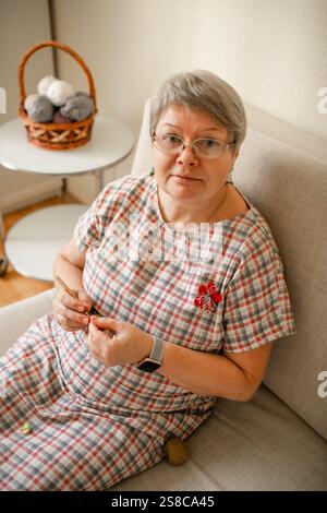 Eine ältere Frau, die zu Hause Brillenhäkeln trägt und auf einem beigefarbenen Sofa sitzt. Stockfoto