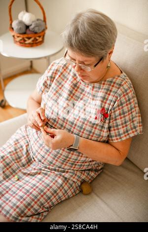 Eine ältere Frau, die zu Hause Brillenhäkeln trägt und auf einem beigefarbenen Sofa sitzt. Stockfoto