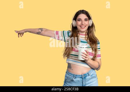 Lächelnde junge Frau mit Tätowierungen, die mit dem Finger zeigen und die wiederverwendbare Kaffeetasse halten, während sie Musik mit Kopfhörern auf gelbem Hintergrund hört Stockfoto