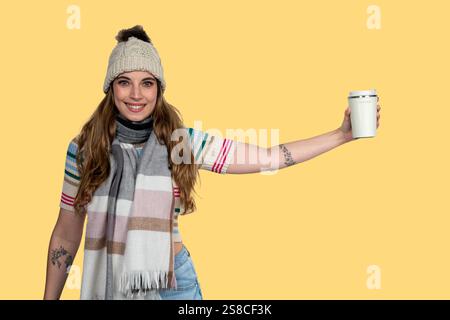 Eine junge Frau in Winterkleidung, die eine wiederverwendbare Kaffeetasse auf gelbem Hintergrund hält Stockfoto