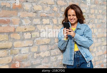 Junge städtische Frau in lässiger Kleidung steht draußen an einer Ziegelwand und schreibt auf ihrem Handy. Stockfoto