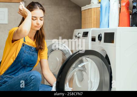 Die müde Frau sitzt auf dem Badezimmerboden und sieht sich schmutziges Geschirr an Stockfoto