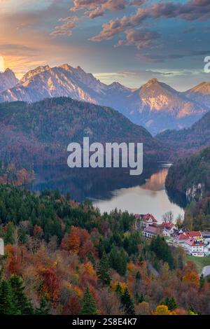 Ein malerischer Blick auf ein Dorf in der Nähe eines ruhigen Sees, umgeben von Herbstlaub und den majestätischen bayerischen Alpen bei Sonnenaufgang, Eine perfekte Mischung aus Stockfoto