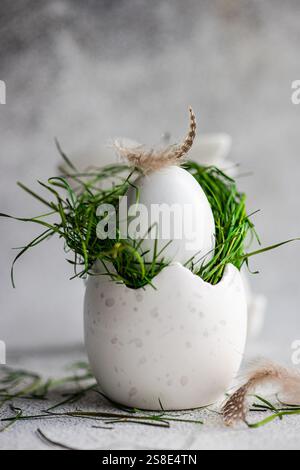 Elegantes und einfaches Osterei mit frischem grünem Gras und zarten Federn auf strukturiertem grauen Hintergrund, das an einen Frühlingsambi erinnert Stockfoto