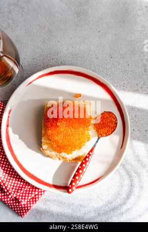 Blick von oben auf einen lebendigen, verführerischen Teller mit rotem Kaviar von Forelle, serviert auf Toast mit einem roten Polka Dot Löffel, vor einem strukturierten grauen Hintergrund Stockfoto