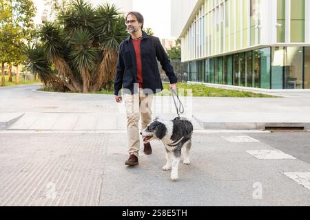 Ein Mann spaziert mit seinem Hund auf einem Fußgängerweg in einer modernen städtischen Umgebung. Das üppige Grün ergänzt die moderne Architektur Stockfoto
