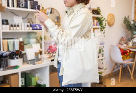 Eine Person untersucht ein Produkt in einem Bio-Geschäft sorgfältig, während sie eine beige Tragetasche mit sich führt, umgeben von umweltfreundlichen Artikeln, die der Shop kreiert Stockfoto