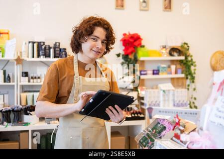 Ein junger Ladenbesitzer mit lockigen Haaren und Schürze verwendet ein Tablet in einem Geschäft. Die Regale sind mit bunten Produkten und Grünpflanzen, Kreationen, gesäumt Stockfoto