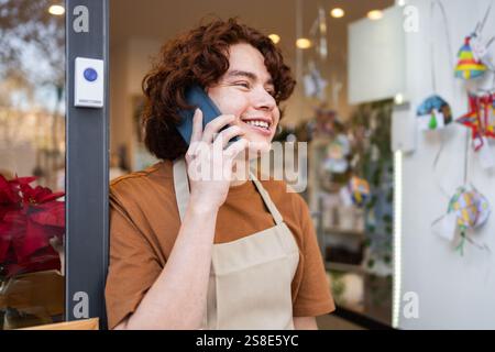 Ein fröhlicher Ladenbesitzer, der eine Schürze trägt, steht am Eingang des Bio-Ladengeschäfts und lächelt, während er ein Telefongespräch im Ladengeschäft führt Stockfoto