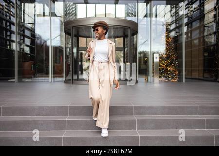 Eine modische schwarze Frau mit einem selbstbewussten Lächeln, in beigefarbenem Outfit und Sonnenbrille, die Stufen vor einem modernen Gebäude Urban Life hinuntergeht Stockfoto